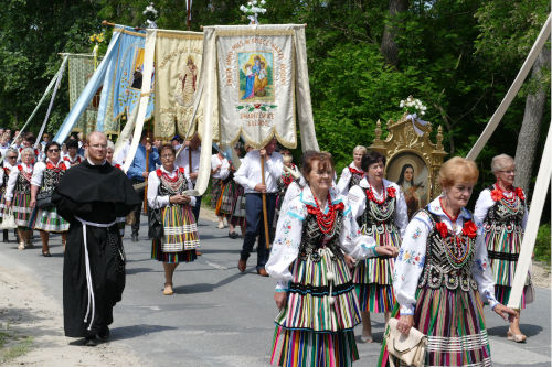 Ludzie idcy w procesji Boego Ciaa - kobiety w ludowych strojach, mczyni nioscy sztandary, obok idcy zakonnik.