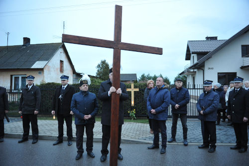 Osoba ubrana w grnicz odwitn czapk, trzymajca duy drewniany Krzy, ktry oparty jest o asfalt na ulicy. Wok niej inne osoby ubrane midzy innymi w mundury straackie.