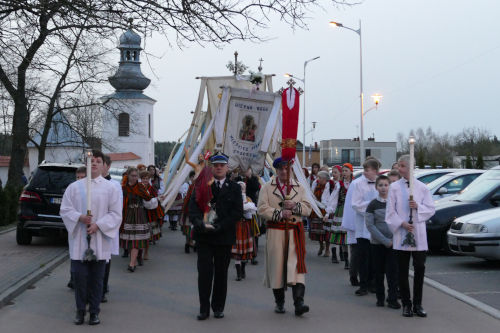 Procesja rezurekcyjna. Idą ludzie ubrani w ludowe stroje, ministranci w komżach. Niektórzy ludzie niosą sztandary.