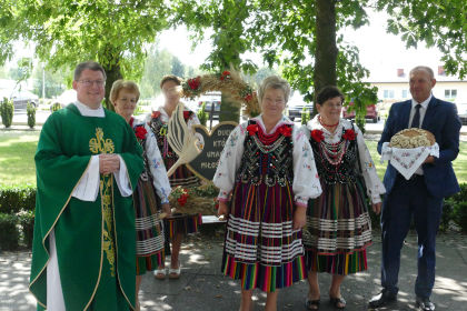 Proboszcz Mariusz Kapczyski stojcy z kobietami w strojach ludowych wraz z doynkowym wiecem. Po prawej stronie stoi mczyzna w garniturze trzymajcy chleb.