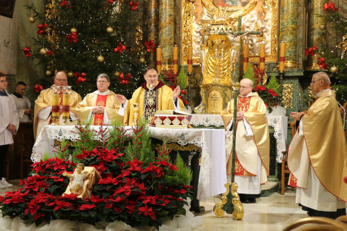 Piciu Ksiy w zotych ornatach celebrujcych Msz wit przy otarzu. Przed Stoem Eucharystyczym znajduje si figurka Jezusa umiejscowiona w czerwonych kwiatach - Gwiazdach betlejemskich. Po lewo w tle choinka oraz ministranci.